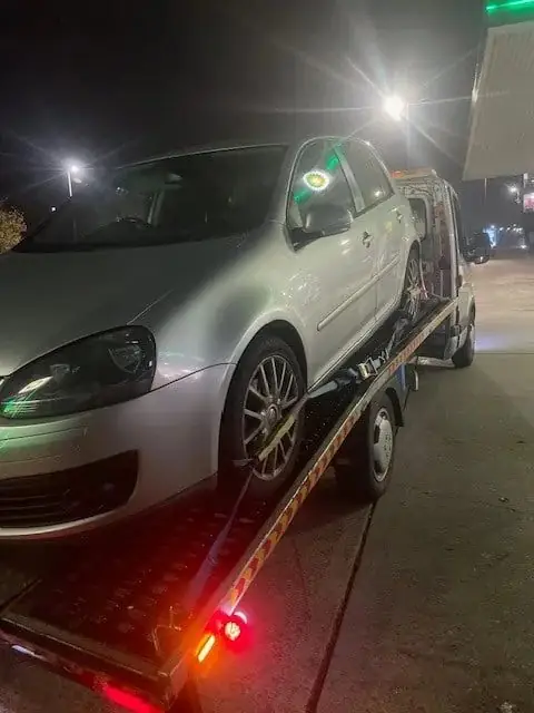 Silver Volkswagen Golf loaded on a recovery truck at a petrol station at night